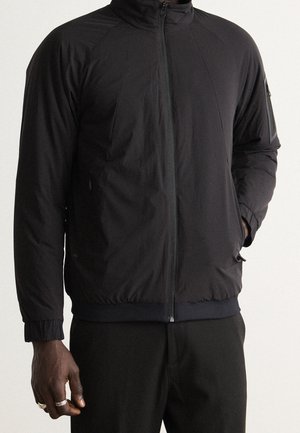 Mężczyzna ubrany w czarną kurtkę zapinaną na zamek z kołnierzem typu stójka, elastycznymi mankietami oraz kieszenią na zamek na górnym lewym rękawie.