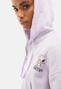 Felpa zip con cappuccio in lavanda, con una texture particolare, caratterizzata da un ricamo di Snoopy sul lato sinistro del petto e un cappuccio con coulisse.