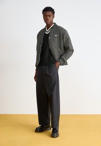 Quilted olive green bomber jacket, black sweater underneath, wide gray trousers, and black platform shoes, set against a yellow background.