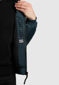 Person som bär svart tröja öppnar mörkgrön vadderad jacka och visar innerficka med Khujo-märkeslapp och detaljerad dragkedja.