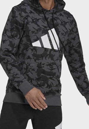 Homme portant un sweat à capuche noir et gris à motif camouflage avec un logo triangulaire blanc et un pantalon noir avec un accent blanc, debout avec une main visible.