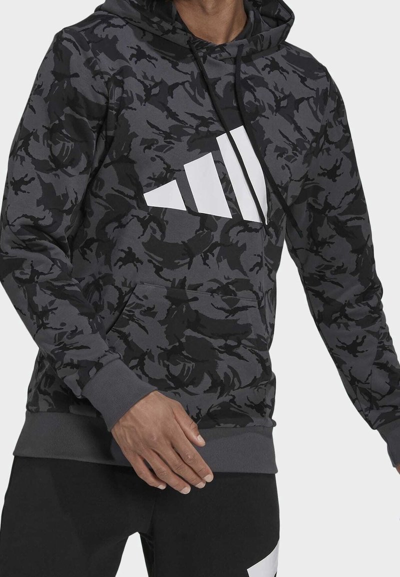Homme portant un sweat à capuche noir et gris à motif camouflage avec un logo triangulaire blanc et un pantalon noir avec un accent blanc, debout avec une main visible.