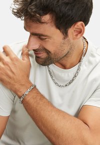 Collier et bracelet en chaîne argentée avec des maillons entrelacés, portés par une personne vêtue d'un t-shirt blanc uni, avec des cheveux courts et foncés.