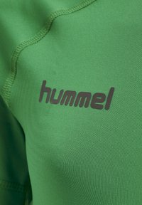 Grøn atletisk stof med en glat tekstur, prydet med et sort "hummel" logo. Sømmen er synlig langs kanterne.