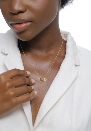 Close-up van een donkerhuidig persoon die een witte blazer draagt en een gouden zonnestraalketting vasthoudt ter hoogte van het sleutelbeen.