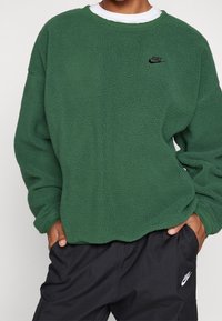 Zelená flísová mikina s texturovaným povrchem a černým logem Nike na hrudi, kombinovaná s černými cargo kalhotami s bílým logem.