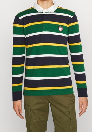 Osoba ubrana w longsleeve typu polo z poziomymi paskami w kolorach zielonym, granatowym, białym i żółtym oraz oliwkowe krótkie spodenki typu cargo.