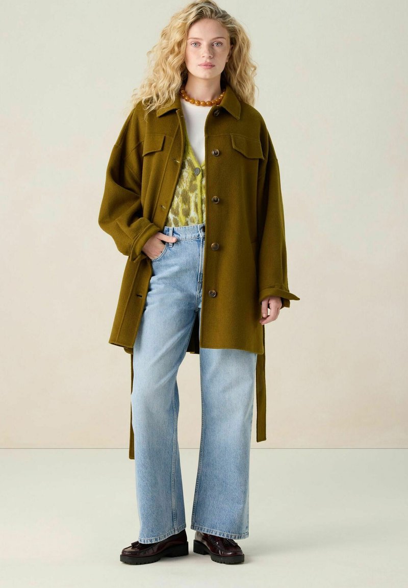 Chemise jacket oversize vert olive avec des boutons sur l'avant et deux poches poitrine, superposée à un pull à motifs et un jean bleu clair. Chaussures marron.