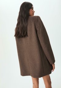 Smeđi oversized vune kaput s ravnim krojem, minimalnim detaljima šivanja i glatkom teksturom, nošen preko svijetlih odjevnih predmeta.