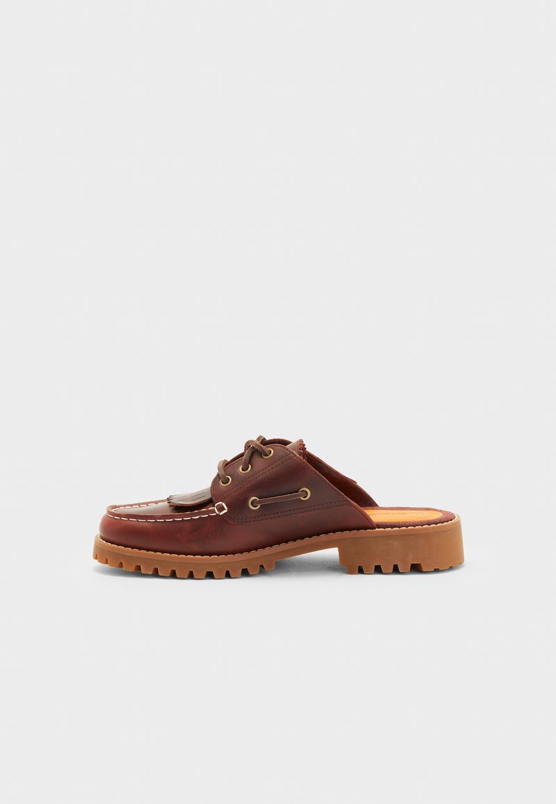 Zapato marrón de cuero tipo mocasín sin cordones, con lazos decorativos, ojales dorados y suela de goma texturizada.