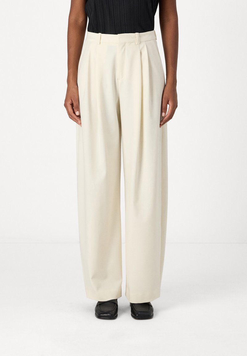 Cream-colored wide-leg trousers with pleats at the waist, made from smooth fabric. Black shoes complete the outfit.