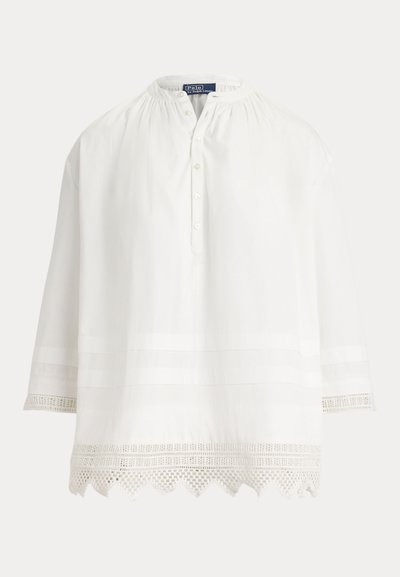 Vit långärmad blus med rund krage, knappar framtill och dekorativ spetskant längs fåll och manschetter.