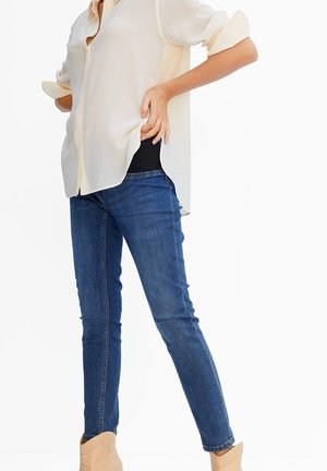 Person iført blå jeans og beige skjorte løfter skjorten for at vise sort undertøjspanel ved taljen, hvilket indikerer graviditets- eller støttetøj.