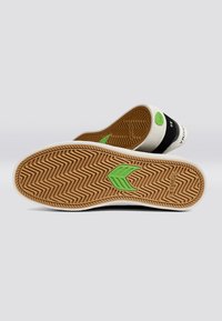 Schwarz-weißes Schuhwerk mit einer gemusterten Gummisohle. Grüner Akzent auf der Sohle und Branding-Text. Strukturiertes Design für besseren Grip.