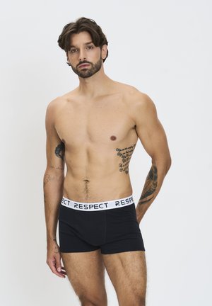 Boxer-briefs noirs avec un waistband élastique blanc portant le mot "RESPECT" répété. Le modèle se tient avec les bras le long du corps et des tatouages visibles.