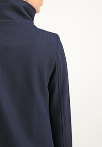 Giacca in misto lana blu navy con colletto alto, caratterizzata da un finitura texturizzata e linee pulite sulle spalle per un look sartoriale.