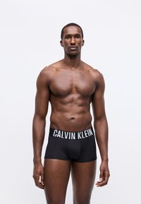 Boxer realizzati in materiale nero con un logo bianco "CALVIN KLEIN" sulla vita. Il tessuto sembra liscia, con un design aderente e cuciture minime.