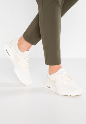 Person wearing light beige Nike sneakers and olive green pants, standing with one foot slightly raised on a white floor and background.