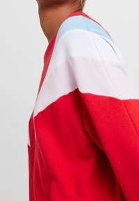 Sweatshirt rouge avec un design coloré en blocs, comprenant des sections blanches et bleu clair, fabriqué en tissu doux, avec des coutures visibles et une coupe décontractée.