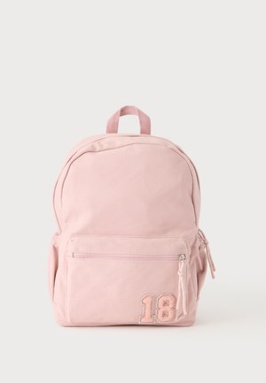 Sac à dos en tissu rose clair avec poche avant zippée et écusson numéro 18 dans le coin inférieur droit, avec poches latérales et poignée supérieure.