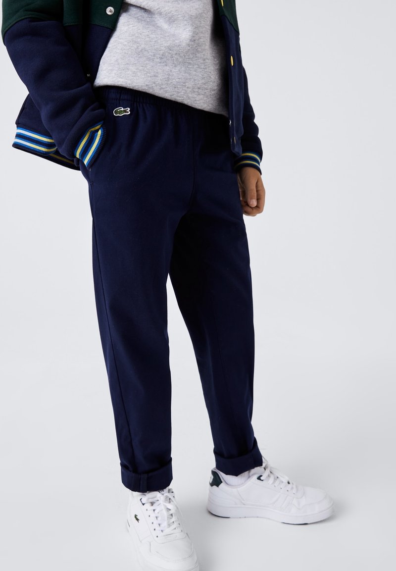 Pantaloni da jogging blu navy con vita elastica, caviglie risvoltate e un piccolo logo verde. Abbinati a scarpe da ginnastica bianche e a un maglione grigio.