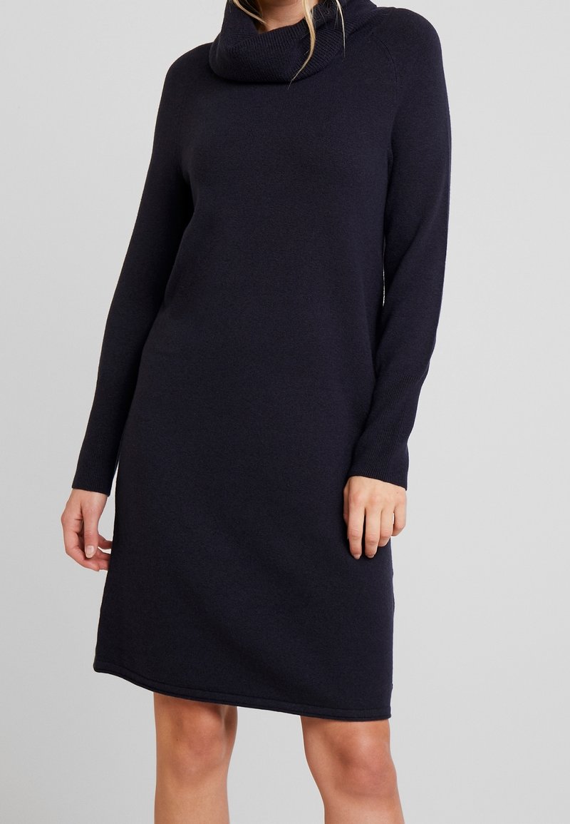Femme portant une robe en maille bleu marine à manches longues avec un col roulé haut et ample sur un fond gris uni.