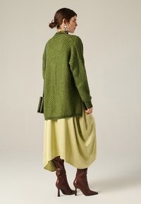 Cardigan in maglia verde con texture, indossato sopra una gonna asimmetrica in seta verde chiaro, abbinato a stivali alti fino al ginocchio marroni con tacco.