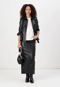 Giacca di pelle nera con chiusura a zip, corpino aderente e due tasche frontali; abbinata a una gonna midi di pelle nera e stivaletti neri.