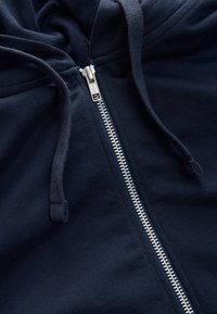 Gros plan d'un sweat à capuche bleu marine montrant une fermeture éclair argentée et des cordons sur un tissu doux.