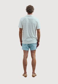 Witte kabelbrei kortemouwenshirt; lichtblauwe zwembroek; lichtbruine geweven instap schoenen; gezien van achter.