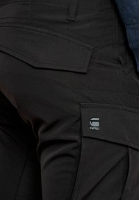 Black fabric cargo pants featuring multiple pockets, contrast stitching, and a grey logo patch on the side. Smooth texture with a fitted silhouette.