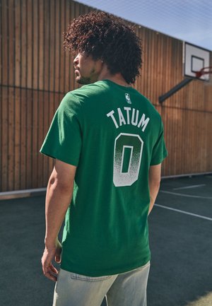 T-shirt en coton vert avec "TATUM" en lettres blanches et "0" au dos. Le modèle a les cheveux bouclés et porte un jean clair. Arrière-plan de terrain de basketball.