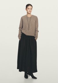 Maglione lavorato a maglia beige con scollo a V, abbinato a una gonna nera maxi e tacchi a punta neri. Presenta maniche lunghe e tasche laterali.