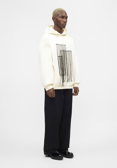 Model z kratkimi blond lasmi nosi belo kapucar z črnim vrvnim vzorcem in besedilom, črne široke hlače in črne čevlje.