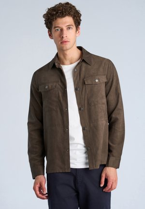 Mann mit lockigem Haar, der eine braune Hemdjacke über einem weißen T-Shirt und dunklen Hosen vor einem einfarbigen hellen Hintergrund trägt.