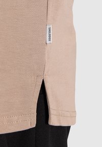 Beige kankaan reuna, jossa on sivuaukko ja pieni valkoinen etiketti, jossa lukee "SMILODOX" mustan tekstuuripintaisen kankaan päällä.