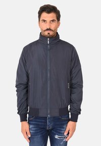 Giacca blu navy scura con colletto alto, zip frontale e polsini elasticizzati. Presenta una texture liscia e tasche laterali, progettata per un look aderente.