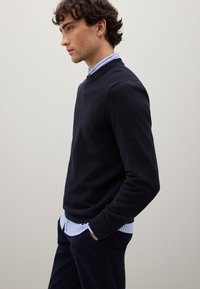 Maglione a maglia strutturata in blu navy indossato sopra una camicia a righe blu chiaro e bianca, abbinato a pantaloni scuri, modello posa di lato contro uno sfondo neutro.