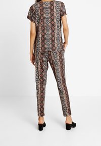 Personne portant un haut et un pantalon à imprimé serpent assortis, debout avec une main dans la poche, mules à talons noirs, fond blanc.