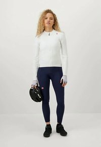 Conjunto de ciclismo que incluye una camiseta ajustada de manga larga blanca, leggings azul marino y zapatos negros. El modelo sostiene un casco, exhibiendo un diseño deportivo.