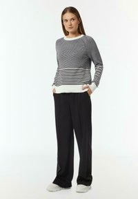 Grau gestreifter Pullover mit ribbeligem weißen Saum, kombiniert mit schwarzen weit geschnittenen Hosen und weißen Sneakers, die seitliche Taschen haben.