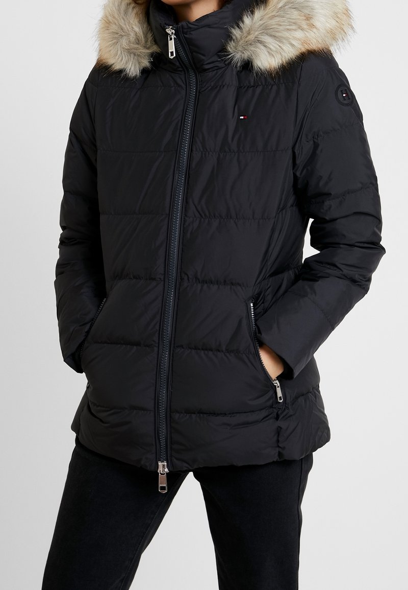 Femme portant une veste d'hiver matelassée noire avec une capuche bordée de fourrure, fermeture éclair à l'avant et mains dans les poches zippées.