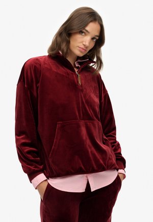 Suéter de terciopelo burdeos con cuello con media cremallera, bolsillo frontal y blusa rosa debajo. Presenta un tejido texturizado y un detalle de logo bordado.