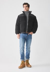 Giovane uomo che indossa una giacca a vento nera, un maglione a collo alto grigio, jeans blu e stivali da lavoro beige, in piedi davanti a uno sfondo bianco semplice.