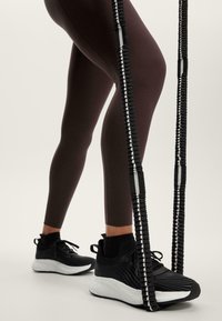 Schwarze Sportschuhe mit strukturiertem Obermaterial und weißen Sohlen, kombiniert mit engen, dunkelbraunen Leggings und einem schwarz-weißen Springseil.