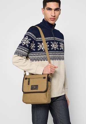 Bolso de mensajero de lona caqui con un detalle de cuero marrón, que cuenta con un bolsillo frontal con cremallera y una etiqueta de cuero con la marca. Incluye una correa ajustable.