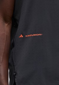 Nahaufnahme eines perforierten schwarzen Stoffs mit orangefarbenem "H.KOUMORI"-Logo und dreieckigem Symbol, neben dem dunkelhäutigen Arm einer Person.