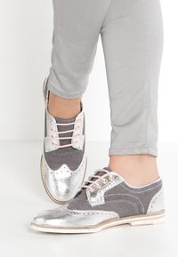 Chaussures à lacets grises et argentées avec un dessus en textile gris texturé, un bout en métal argenté brillant et des lacets roses ; ornées d'accents perforés.