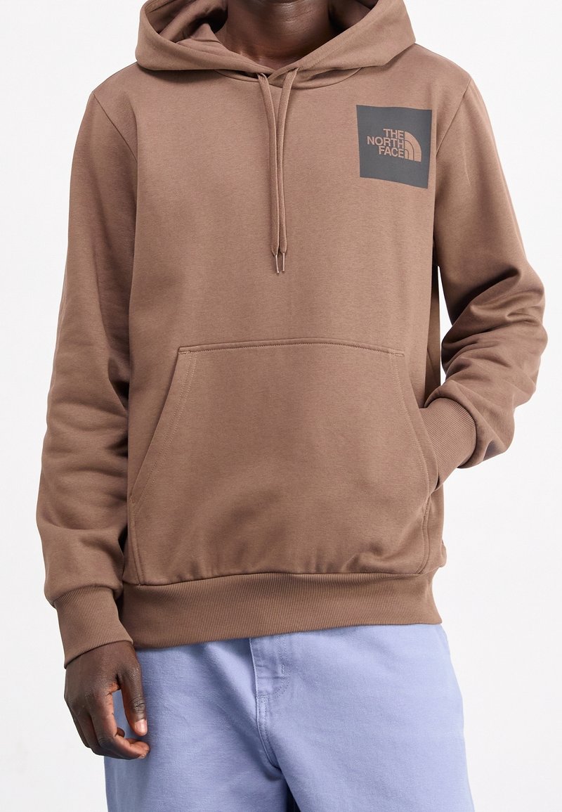 Homme portant un sweat à capuche marron The North Face avec poche avant et cordons, associé à un pantalon bleu clair, main dans la poche.