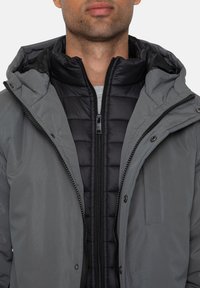 Grau isolierte Jacke mit einem schwarzen Reißverschluss-Innenfutter, gepolstertem Design, abnehmbarem Kapuzen und Druckknopfverschlüssen an der Vorderseite.